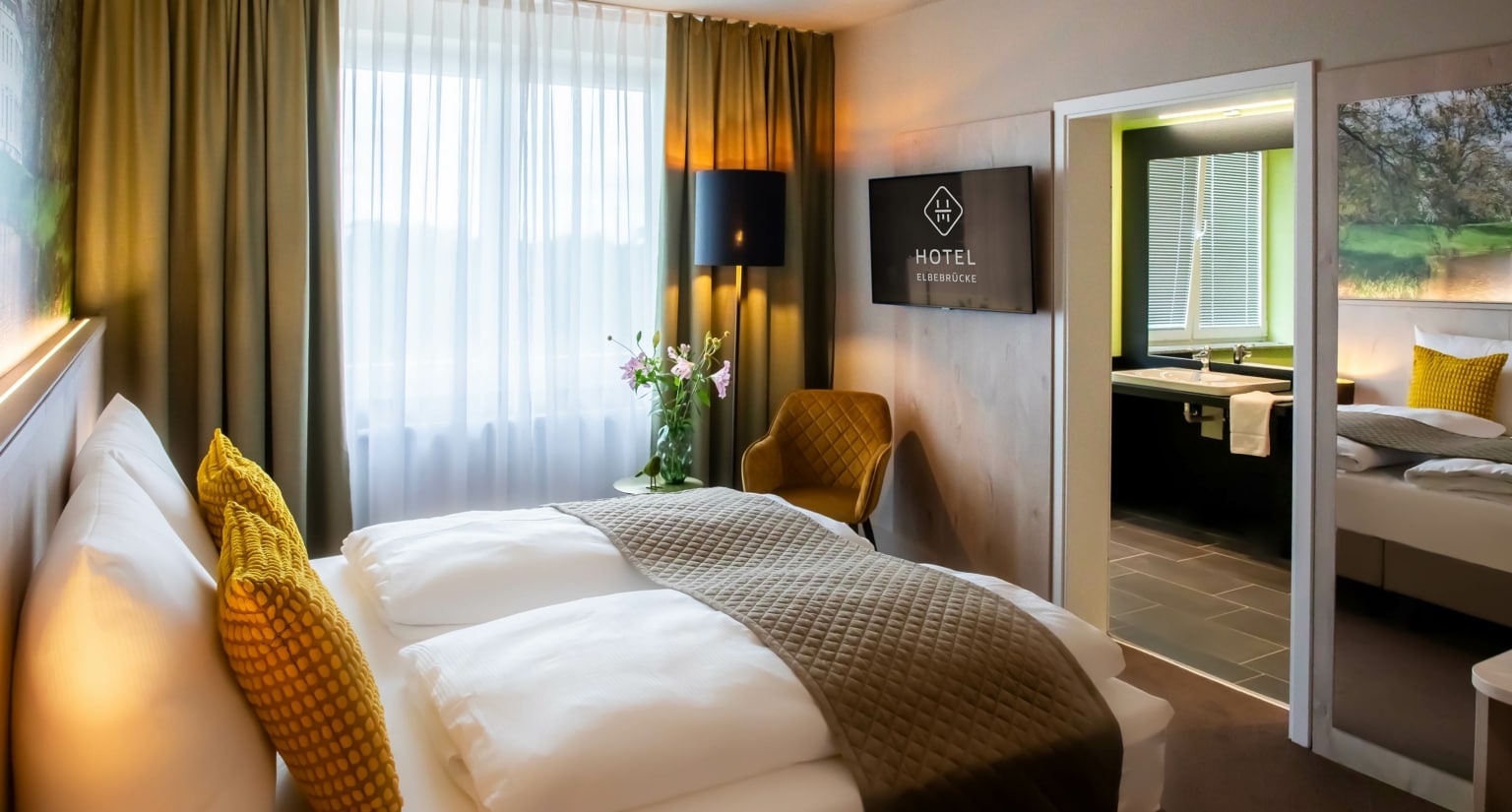 Aufnahme eines Hotel-Doppelbettes mit separatem Badezimmer, bodentiefen Fenstern und TV, welches zum Übernachten bei Dessau einlädt