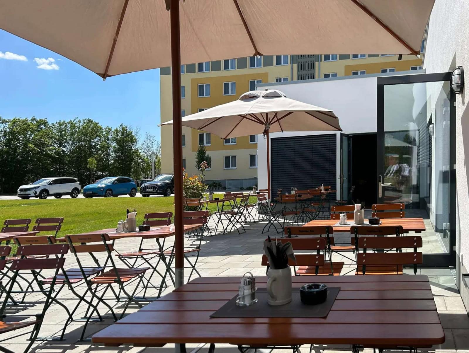 Foto des Restaurant-Außenbereiches mit Holztischen und Stühlen, im Hintergrund sind drei Autos und ein Hotelgebäude zu sehen