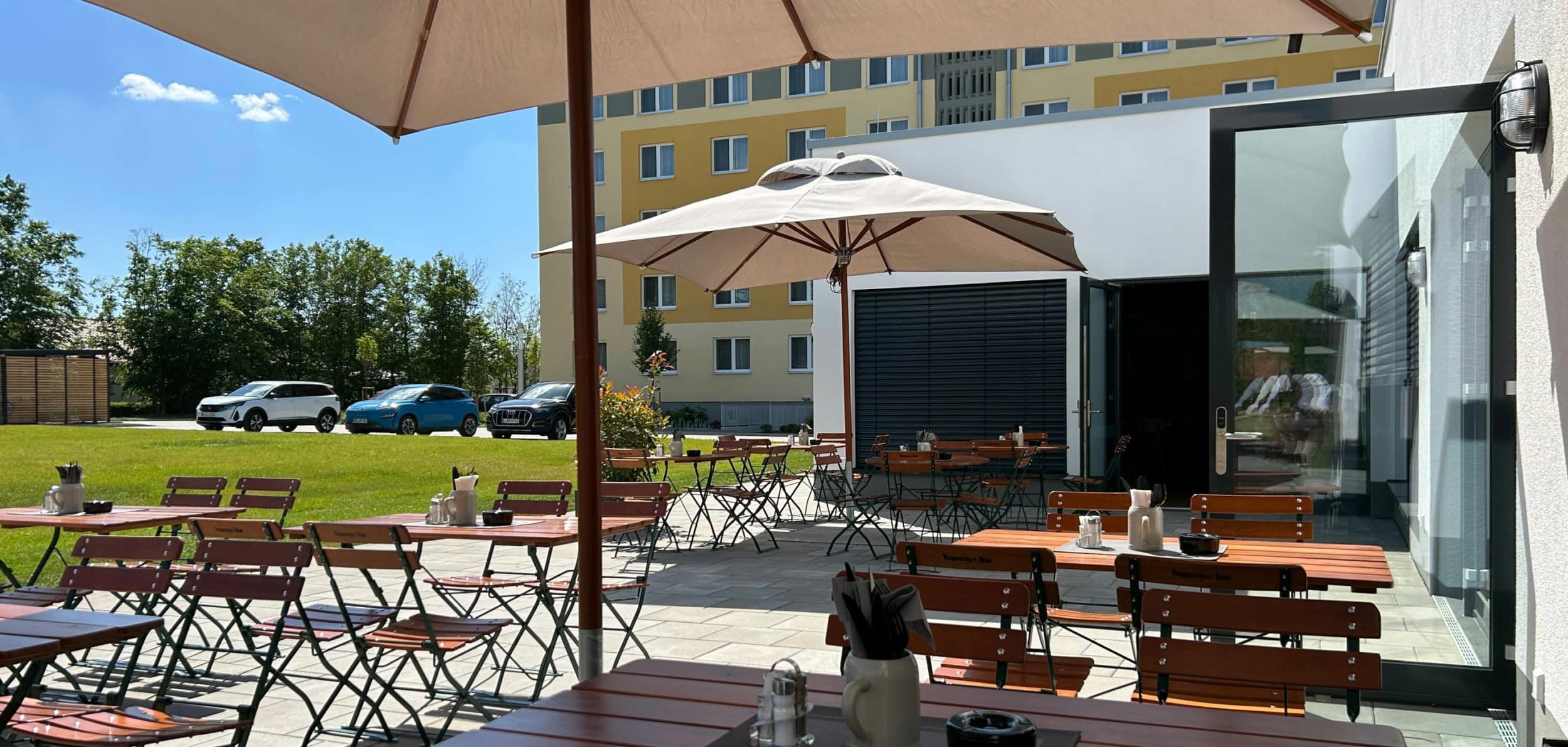 Foto des Restaurant-Außenbereiches mit Holztischen und Stühlen, im Hintergrund sind drei Autos und ein Hotelgebäude zu sehen