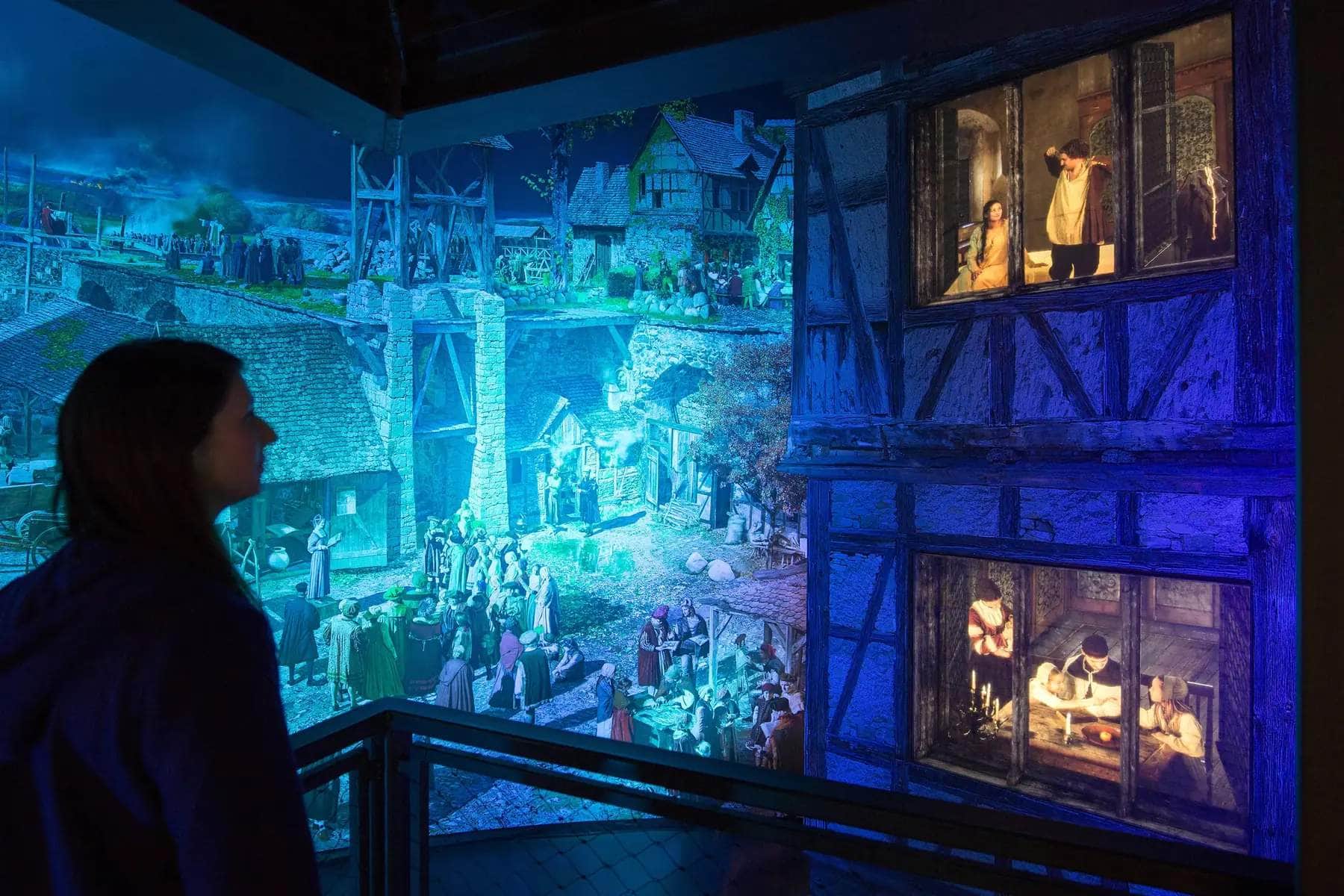 Eine Frau schaut auf eine bläuliche Lichtprojektion, die Alltagsszenen aus einem mittelalterlichen Haus zeigt