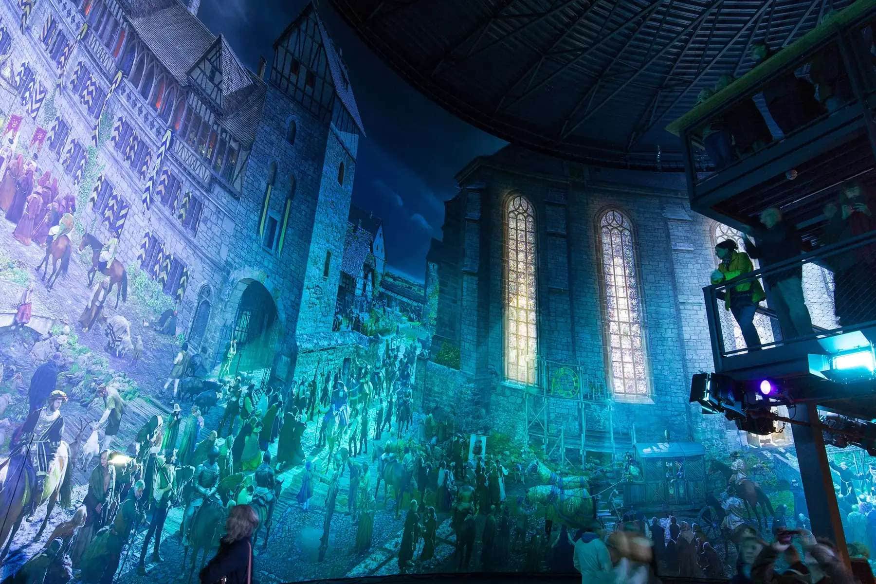 Mehrere Besucher des Asisi Panoramas in Wittenberg stehen auf Aussichtsplattformen und betrachten eine 360 Grad Lichtprojektion