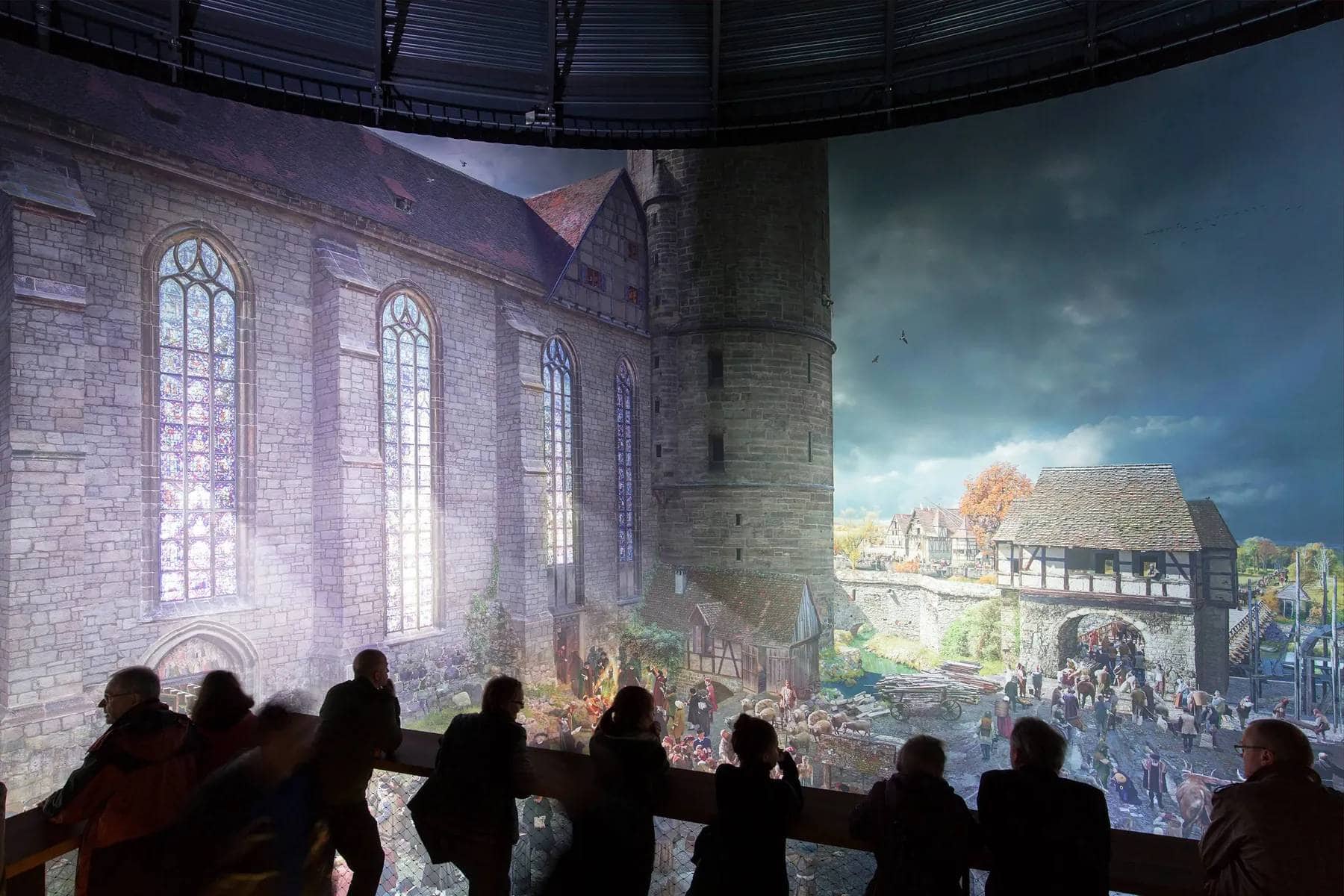 Personen auf einer Holzbalustrade bestaunen eine Lichtprojektion, die die Schlosskirche Wittenbergs im Jahre 1517 zeigt