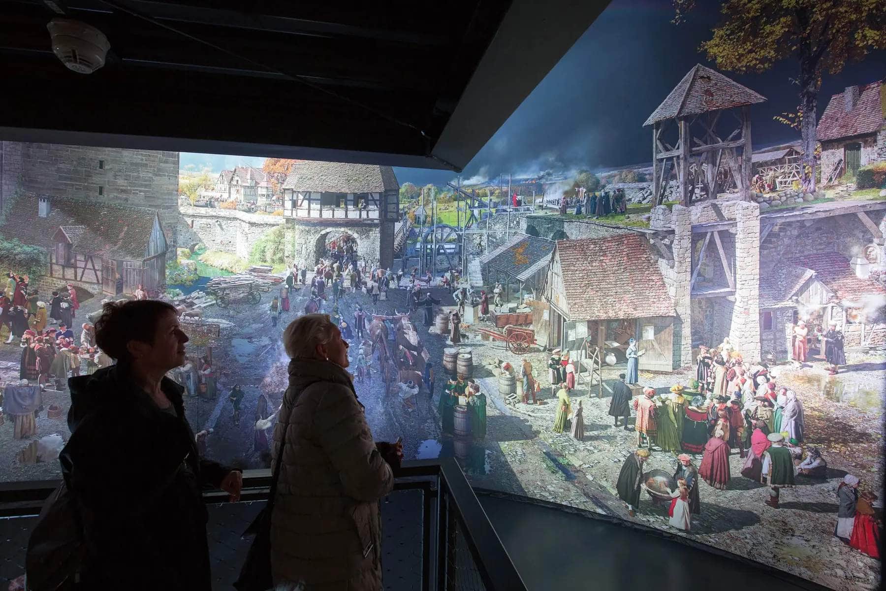 Zwei ältere Damen schauen auf eine Lichtprojektion, die verschiedene Szenen in einer Mittelalter-Stadt zeigt